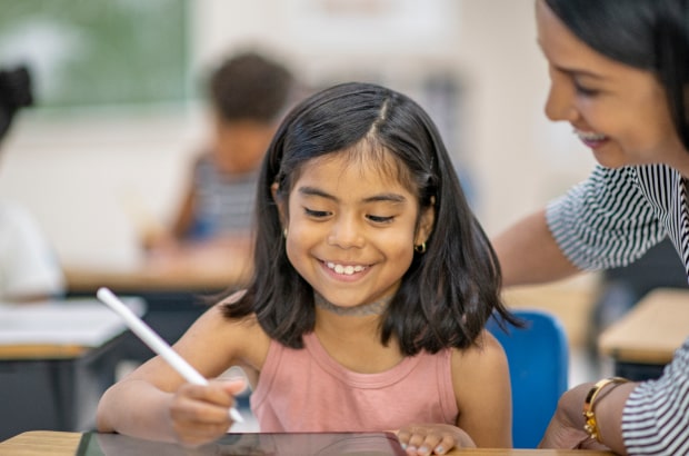 Professora e aluna sentadas à mesa olham juntas a tela de um tablet.