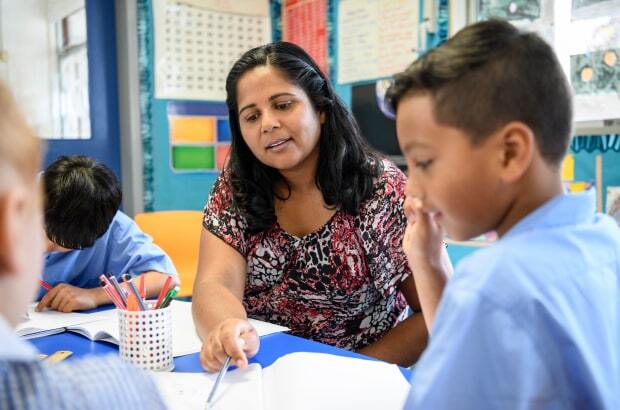 Professora sorri e aponta caderno do aluno, que está sentado, escrevendo.