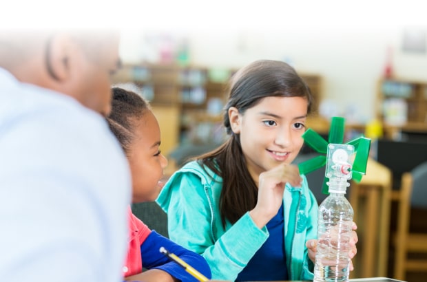 Duas alunas estão sentadas manuseando um cata-vento de plástico, posicionado sobre uma classe. Ao lado delas aparece o perfil do professor, de costas para a câmera.