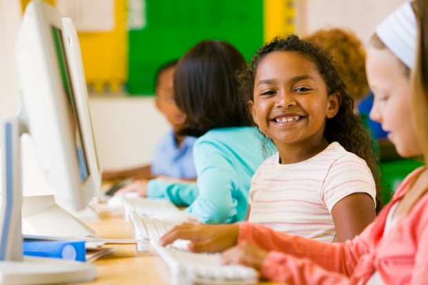 Aluna sorri enquanto trabalha com um computador ao lado de outros estudantes.