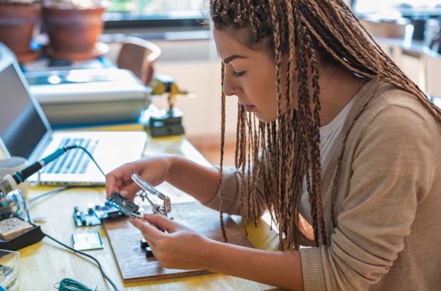 Jovem aluna manipula uma placa de prototipagem em uma bancada, onde também está disposto um laptop.