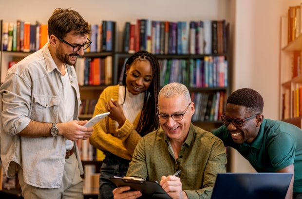 Quatro professores, três homens e uma mulher, estão reunidos em uma biblioteca, ao redor de uma mesa (um deles sentado), conversando e sorrindo. Dois deles manuseiam dispositivos digitais.