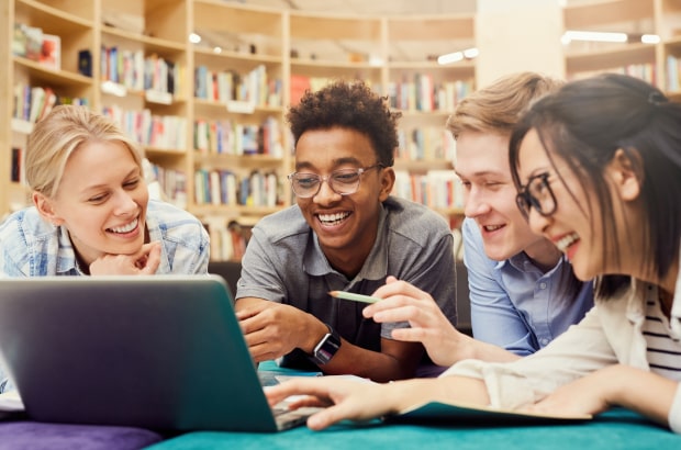 Quatro jovens apoiados sobre uma mesa sorriem enquanto manuseiam um laptop. Um deles aponta para a tela com um lápis.