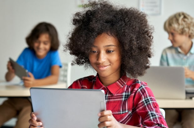 Três alunos aparecem sentados em uma sala de aula manuseando tablets (duas meninas e um menino). Em primeiro plano, uma das meninas olha para seu tablet e sorri.