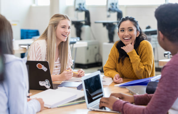 Quatro alunos aparecem sentados ao redor de uma mesa, em frente a um laptop, e as duas meninas que encontram-se de frente, sorriem.